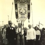 Gruppe von Menschen. Ein Mann trägt ein Banner mit Aufschrift "Untergruppe Schöneberg - Kolonie Lindenhain. 1913 - 1945